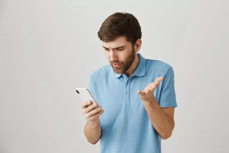 Bearded young Caucasian man wearing a light blue shirt looking at his mobile phone and showing signs of frustration by cookie_studio on Freepik.