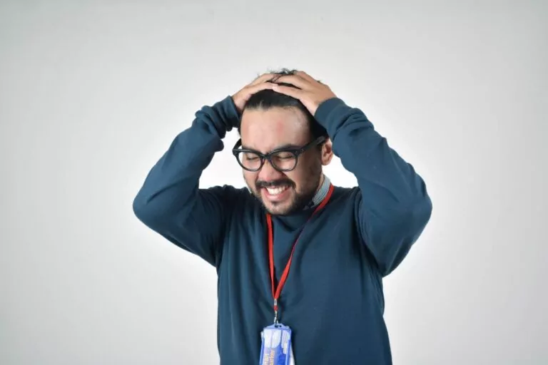 A frustrated male office worker with a short beard wearing eyeglasses, office ID on a red lanyard, and dark teal longsleeved shirt showing extreme frustration and holding his head with both hands.