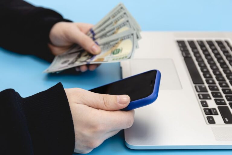 Close-up photo of hands with wrist-length black sleeves holding money on the left and a smartphone on the right in front of a laptop on a light blue background by pvproductions on Freepik.