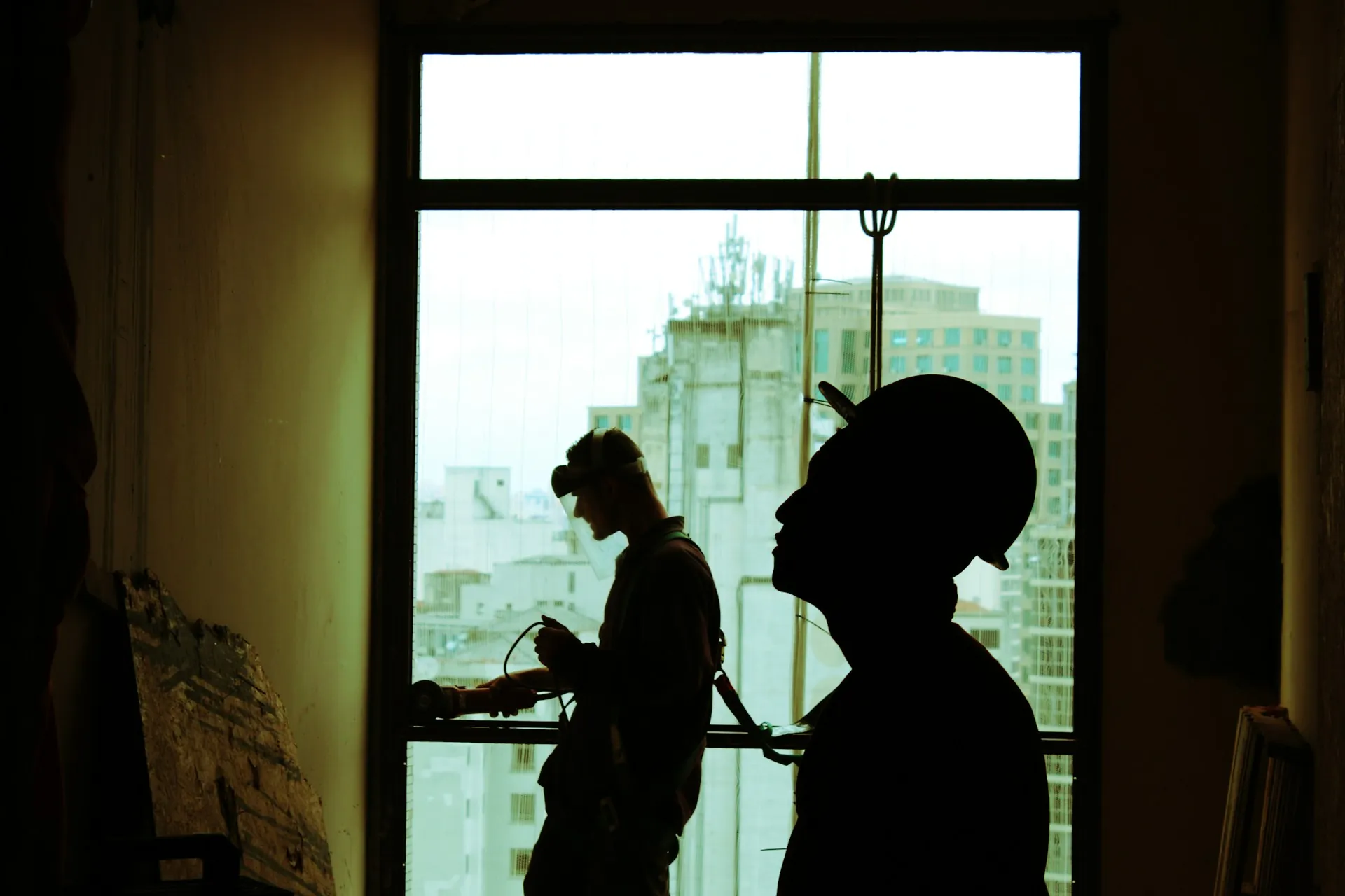 Two construction workers working on site, pictured against the light from a window.