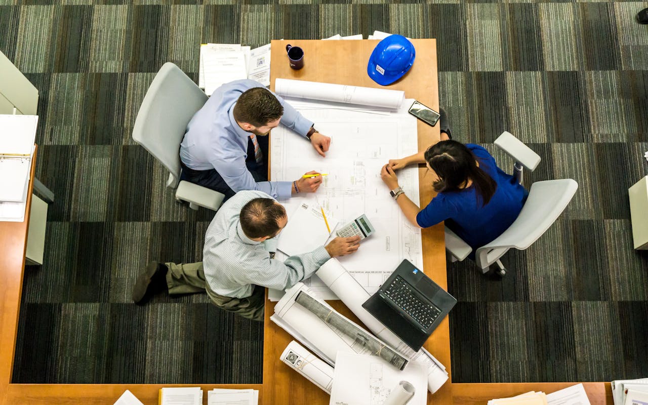 Uma reunião entre funcionários de construção, falando sobre um plano de projeto em uma mesa. Implemente um software de controle de tempo para construção.