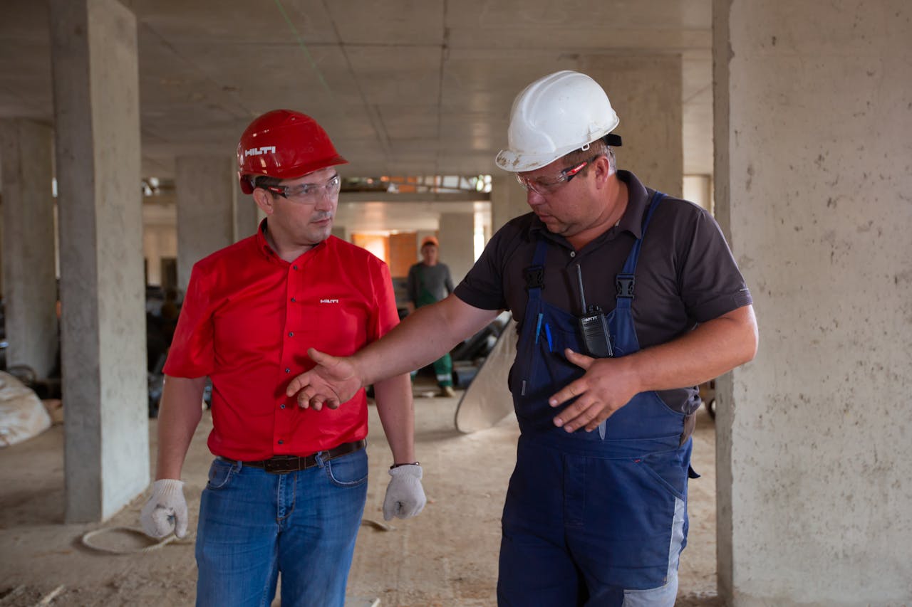 Dois pedreiros conversando, um usando um capacete de segurança vermelho e o outro usando um branco. Controle de horas de obras espalhadas.