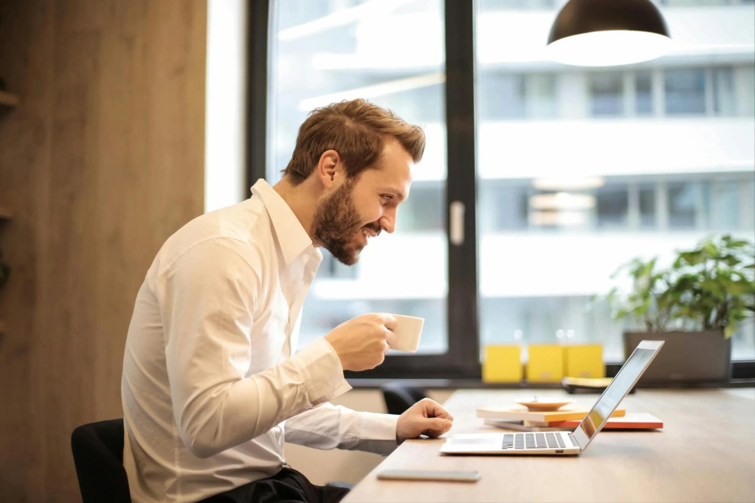 Man smiling while working on a laptop in a bright office, holding a coffee cup.