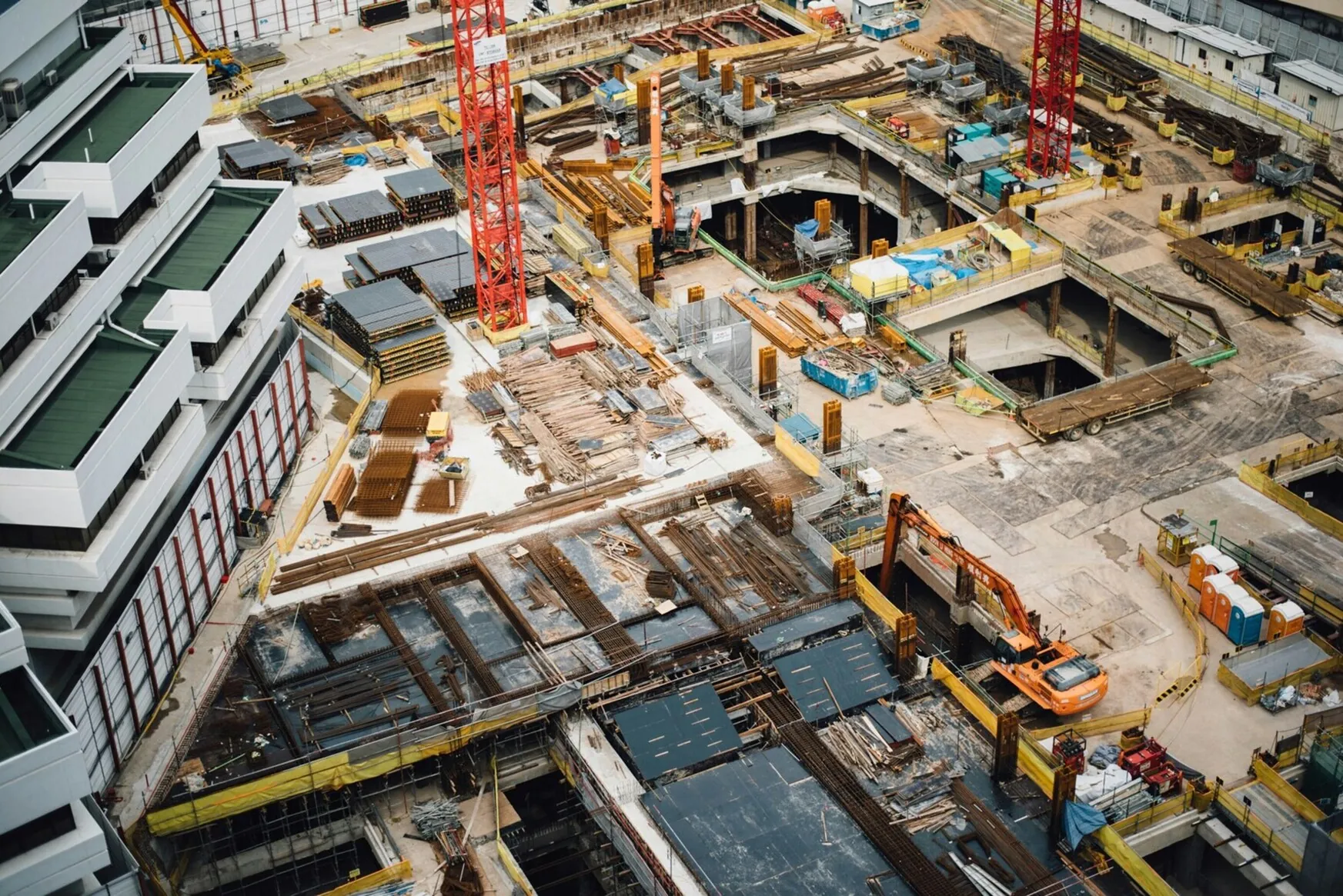 An aerial view of a busy construction site with cranes, heavy machinery, building materials, and partially constructed structures, showcasing an urban development project in progress.